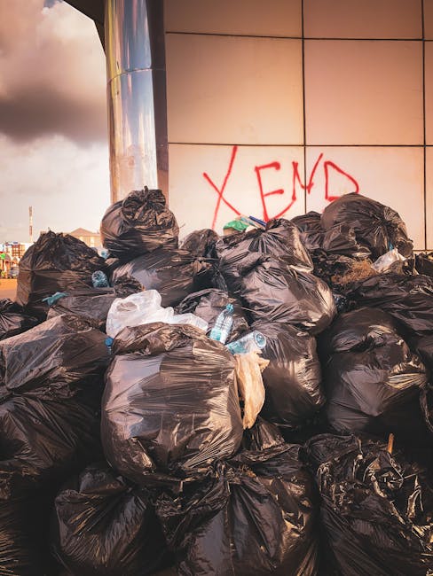 Three large black plastic garbage bags filled with waste are placed on the edge of a paved street, near a curb. The bags appear to be tightly packed and slightly crumpled, with some branding or text v