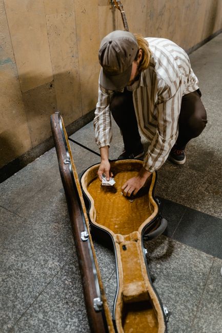 A person wearing a grey cap, plaid shirt, and dark pants is crouching down on a tiled floor inside a building, placing paper money into a large, open, cream-colored guitar case that is lying flat on t