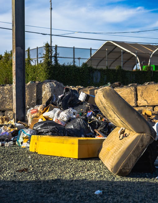 A large pile of mixed rubbish and waste items accumulated on a paved area in front of a shopping centre during daytime. The pile includes various cardboard boxes, plastic bags, paper waste, and discar